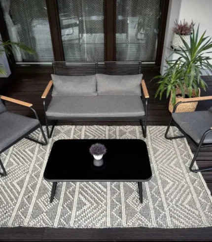 Zestaw ogrodowy RIO widziany z góry: sofa dwuosobowa, dwa fotele z czarnym technorattanem i podłokietnikami polywood oraz stolik kawowy ze szklanym blatem – elegancko ułożone na dużym dywanie na drewnianym tarasie